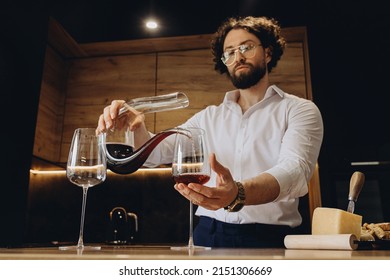 Handsome Curly Man At Home In A Beautiful Kitchen Pours Red Wine Into A Glass With Decanters. A Sommelier Pouring Red Wine Into Decanter