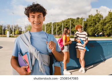 Handsome curly teen high school boy guy with braces bag in casual clothes holding netbooks copybooks smiling looking at camera spending time with friends classmates. Students teenagers hanging out - Powered by Shutterstock