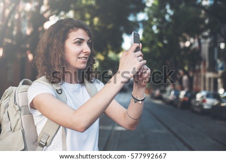 Handsome curly woman taking photo with mobile phone. Walking along the street with backpack and traveling, tattoo on the arm