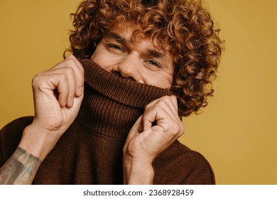 Handsome curly young man hiding half face behind a sweater neck against yellow background