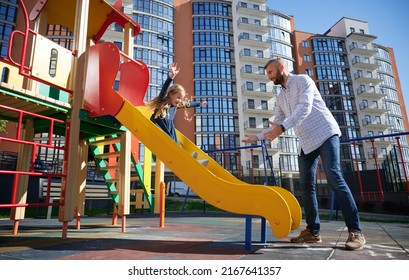 Handsome dad catching cheerful daughter, riding on slide at modern courtyard of city residential high-rise buildings. Bearded father catching pretty girl while standing near slide on playground. - Powered by Shutterstock