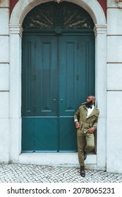 A handsome dapper adult bald bearded man with a cigar in his mouth and in an elegant green overalls costume with a bow-tie is leaning against the wall next to the huge portal doors of an antique house