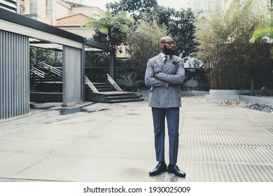 A handsome dapper hairless adult black man entrepreneur in elegant eyeglasses and a fashionable custom-made costume, with a well-groomed beard, is standing outdoors on a metallic floor, looking aside