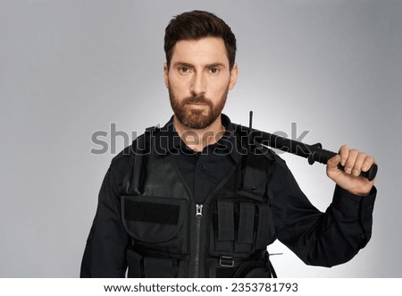 Handsome dark-haired police officer carrying baton on shoulder in studio. Portrait of male policeman with beard, posing to camera with rubber bludgeon, on gray background. Work, profession concept.