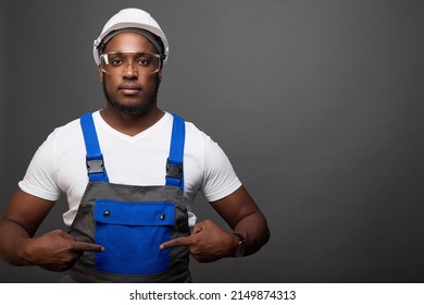 Handsome dark-skinned man gestures to the blue pocket of his new work overalls. Stylish modern worker tries on a new uniform and protective accessories issued in stock. Place for a layout or mock-up