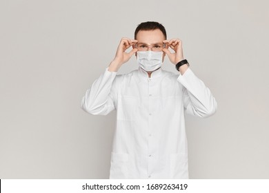 Handsome Dentist Or Doctor Wears A White Medical Gown, Face Mask And Protective Glasses On Grey Background. Concept Of Healthcare And Stomatology