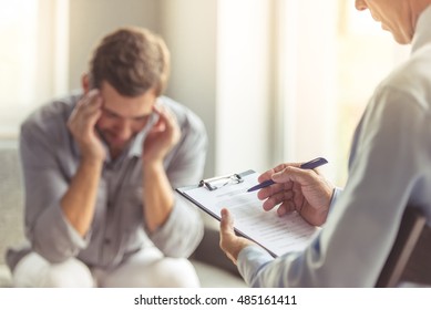 Handsome depressed man in casual clothes is leaning on his knees and telling about his problems while sitting on the couch at the psychotherapist. Doctor in focus - Shutterstock ID 485161411