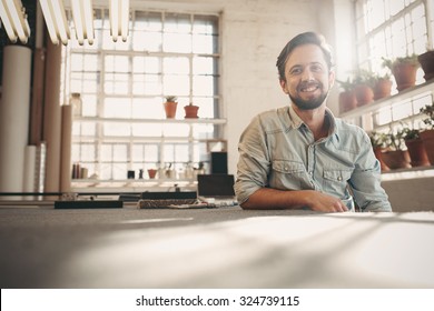 Handsome Designer Entrepreneur Smiling At The Camera While Relaxing In His Studio With Gentle Sun Flare Coming In Through The Window