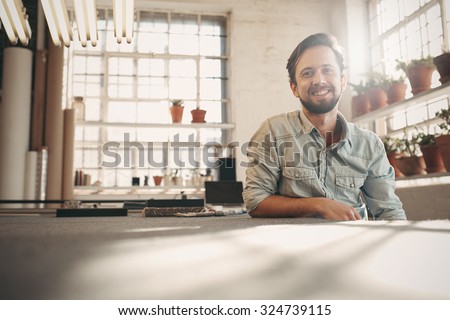 Handsome designer entrepreneur smiling at the camera while relaxing in his studio with gentle sun flare coming in through the window