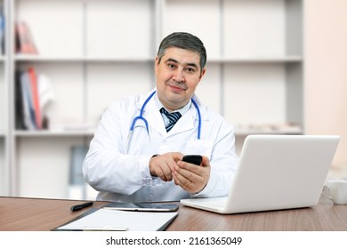 A handsome doctor man at his desk holds a cellphone in his hands and looks at the camera smiling. Modern doctor in the clinic. - Powered by Shutterstock