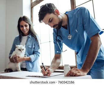 Handsome Doctor Veterinarian At Vet Clinic Is Examining Cute Cat While His Owner Is Standing Nearby And Holding Pet On Hands.