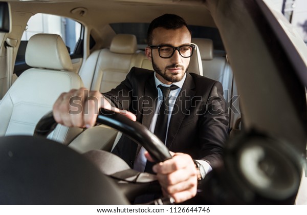 handsome driver in suit driving car and looking at\
side mirror
