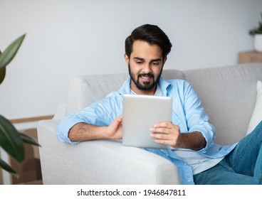 Handsome Eastern Man Browsing Internet On Digital Tablet While Relaxing At Home. Young Smiling Arab Guy Sitting On Couch In Living Room Using Tab Computer, Shopping Online Or Surfing Web, Free Space