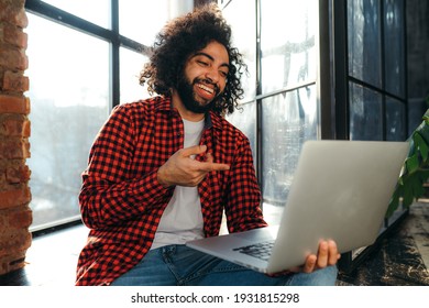 Handsome egyptian student in red plaid shirt holding a computer and pointing at it with a finger while laughing