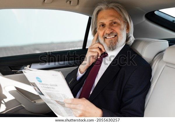 A handsome elderly businessman is driving in\
the car and reading\
newspapers