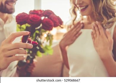 Handsome elegant guy is proposing to his beautiful girlfriend, giving her roses and smiling while they having a romantic date at home