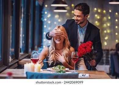 Handsome elegant man is holding roses and covering his girlfriend's eyes while making a surprise in restaurant, both are smiling - Powered by Shutterstock