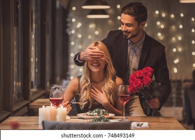 Ein hübscher, eleganter Mann hält Rosen und bedeckt die Augen seiner Freundin, während eine Überraschung im Restaurant, beide lächeln