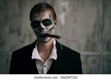 Handsome elegant man with skull makeup poses on a grunge background with a cigar in his mouth sinisterly smiling. Dia de los muertos. Day of The Dead. Halloween.