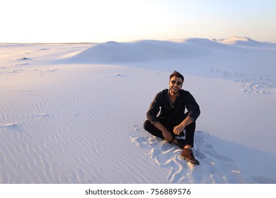 Handsome Emirate man sits on white sand of desert, possing and smiles broad smile, looks around neighborhood on clear evening. Swarthy Muslim with short dark hair and sunglasses dressed in black