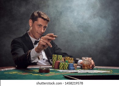 Handsome emotional man is playing poker sitting at the table in casino against a white spotlight.