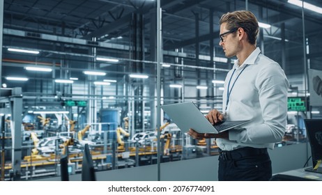 Handsome Engineer In Glasses And White Shirt Using Laptop Computer And Looking Out Of The Office At A Car Assembly Plant. Industrial Specialist Working On Vehicle Design In Technological Facility.