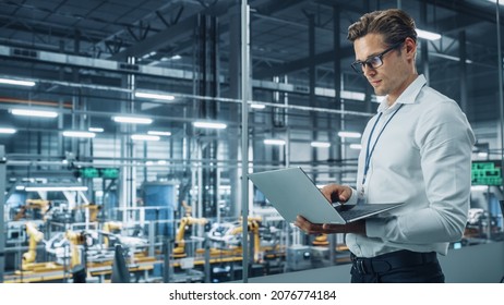 Handsome Engineer In Glasses And White Shirt Using Laptop Computer And Looking Out Of The Office At A Car Assembly Plant. Industrial Specialist Working On Vehicle Design In Technological Facility.