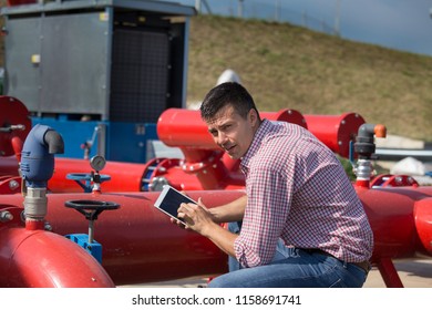 Handsome engineer with tablet squatting in front of pipeline at outdoor industry plant