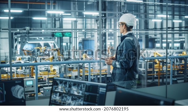 Handsome Engineer in Uniform and Hard Hat Using
Tablet Computer at a Car Assembly Plant. Industrial Specialist
Working on Vehicle Design, Overlooking Production in Technological
Facility.