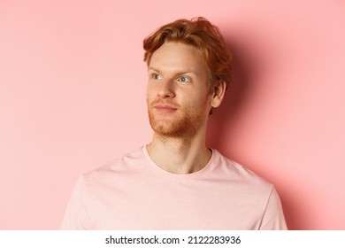 Handsome European Male Model With Red Hair And Beard, Turn Head And Looking Pleased At Copy Space On Left Side, Standing Over Pink Background