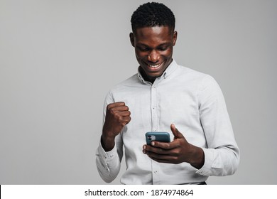 Handsome excited young african business man using mobile phone isolated over gray background, wearing white formal shirt, celebrating success