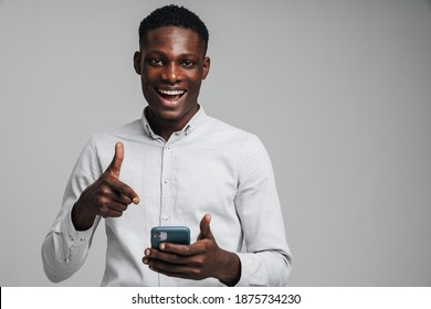 Handsome Excited Young African Business Man Pointing Finger At Mobile Phone Isolated Over Gray Background, Wearing White Formal Shirt