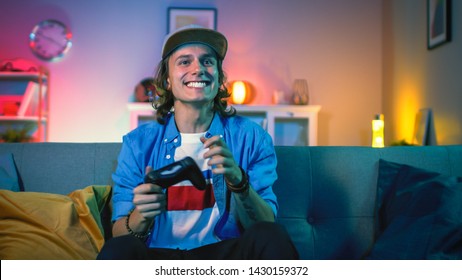 Handsome Excited Young Gamer with Long Hair and a Cap is Sitting on a Couch and Playing Video Games on a Console. He Plays with a Wireless Controller. Cozy Room is Lit with Warm and Neon Light.
