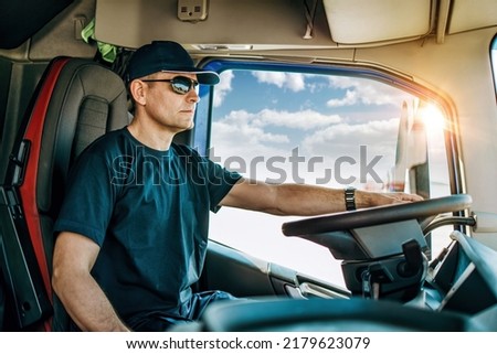 Handsome experienced male truck driver with a hat sitting and driving his truck. Professional transportation and truck drivers concept. Side view.