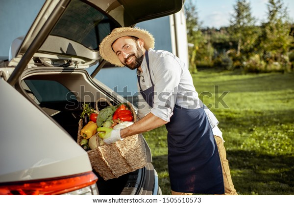 Handsome farmer in apron and straw hat putting a\
basket full of freshly picked vegetables into the car trunk at a\
country cottage