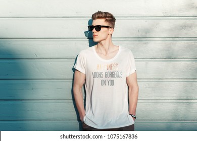 Handsome Fashion Man Hipster In Stylish Sunglasses In A White T-shirt Posing Near A Blue Wooden Wall On The Beach On A Sunny Day. Happiness Looks Gorgeous On You