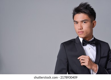 Handsome Fashion Model.
Elegant Man Wear Formal Black Suit With Bow Tie On Gray Background 

