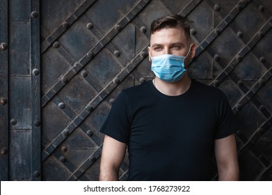 Handsome Fashionable Young Man In A Black T-shirt And A Disposable Medical Mask On A Dark Background. Caucasian Man Correctly Put On Face Mask.