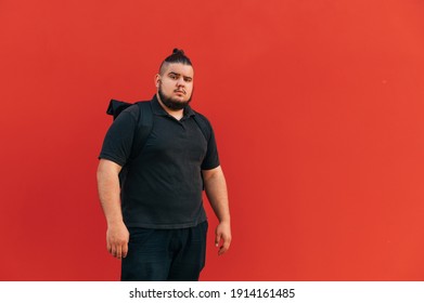 Handsome Fat Hispanic Man With Backpack On His Back Isolated On Red Background, Looking At Camera With Serious Face, Wearing Black Clothes. Copy Space