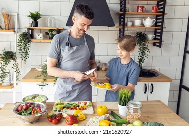 Handsome Father And His Teenager Son Spending Quality Time Together. Men Doing Chores, Cooking Healthy Vegetable Salad, Tasty Food In The Kitchen At Home