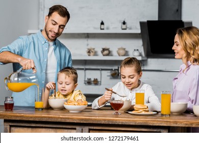 Handsome Father Pouring Orange Juice In Glass While Children Eating Oatmeal At Table