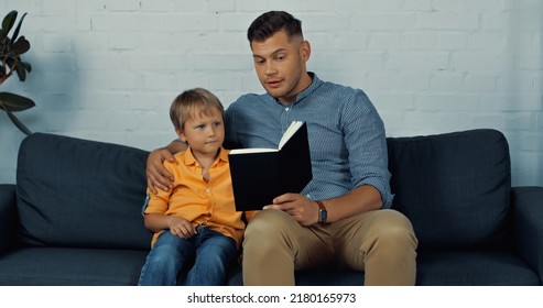 Handsome Father Reading Book Aloud To Son In Living Room