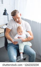 handsome father sitting on chair, talking on smartphone, looking at notebook and holding adorable baby daughter at home - Shutterstock ID 1262323234