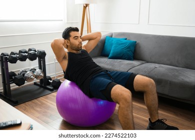 Handsome fit man doing sit-up exercises using a stability ball while working out in his living room home - Powered by Shutterstock