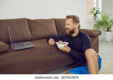 Handsome fit man using laptop after active indoor weightloss fitness workout. Smiling guy sitting on floor by sofa in living room, holding bowl, eating healthy food, watching movie, restoring energy - Powered by Shutterstock