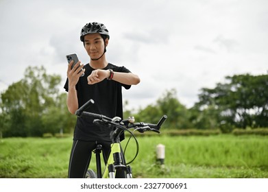 A handsome and fit young Asian man in sportswear and a bike helmet is on his bike, using his smartphone and looking at his smartwatch. - Powered by Shutterstock