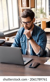 Handsome Focused Man Working Using Laptop Sitting At Office Desk Table In Coworking Workspace. Businessman Freelancer, Executive Manager Or Employee Making Report, Searching Information On Internet