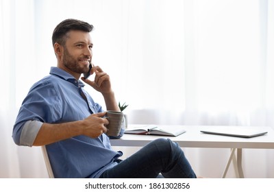 Handsome freelancer man talking on cellphone and drinking coffee at home, millennial guy sitting at desk with laptop and notepad near window, enjoying mobile communication and remote job, copy space - Powered by Shutterstock