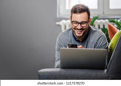 Handsome Freelancer Working From Home, Lying On His Stomach On The Couch With Laptop And Phone. Smiling. Wearing Glasses. Copy Space. 