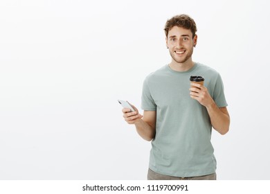 Handsome Friendly-looking Mature Guy With Fair Hair, Holding Smartphone And Cup Of Coffee, Smiling Broadly, Drinking Beverage And Hanging Out With Friends In Park, Standing Carefree Over Grey Wall